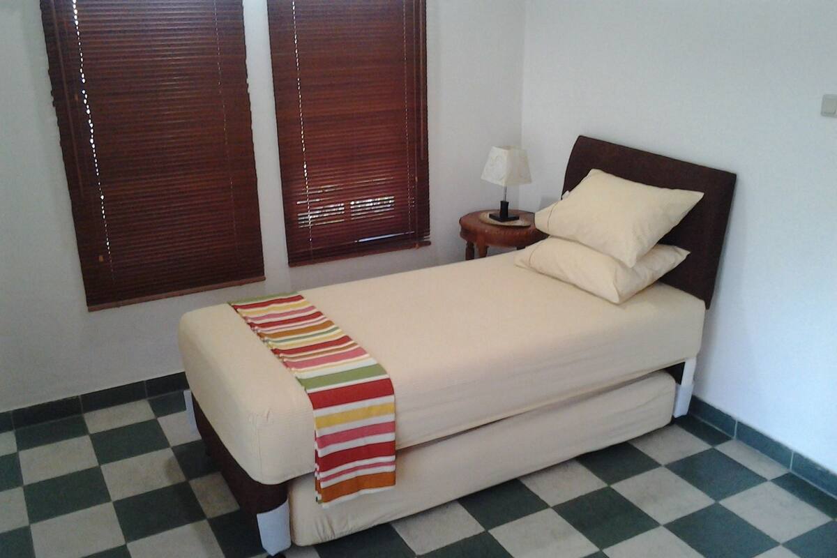 Uma cama de solteiro com um cobertor listrado, um abajur e duas janelas com persianas.
