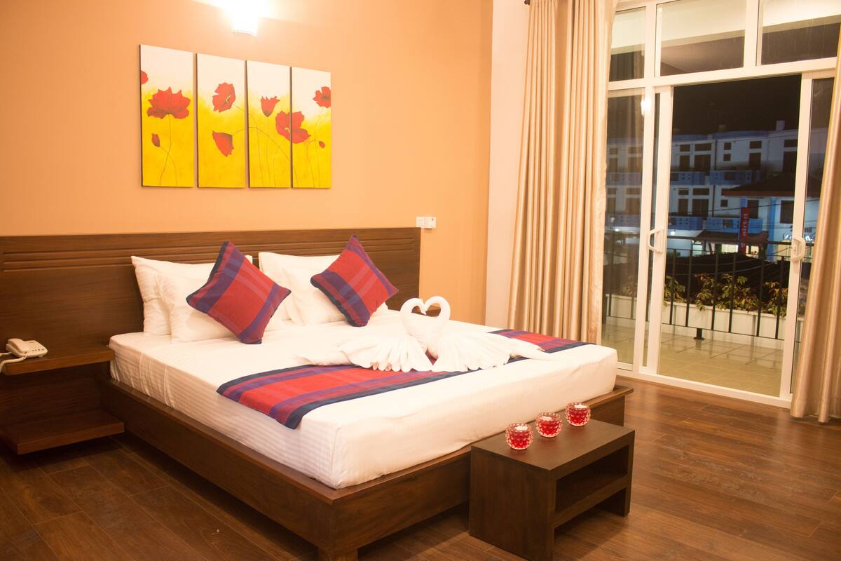 Una habitación de hotel con una cama grande, una mesita de noche, una mesilla con dos objetos decorativos rojos y una ventana que ofrece vistas a edificios y un balcón.