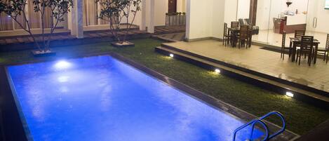 Una piscina de color azul, rodeada de un pequeño jardín con árboles y un patio amoblado al aire libre.