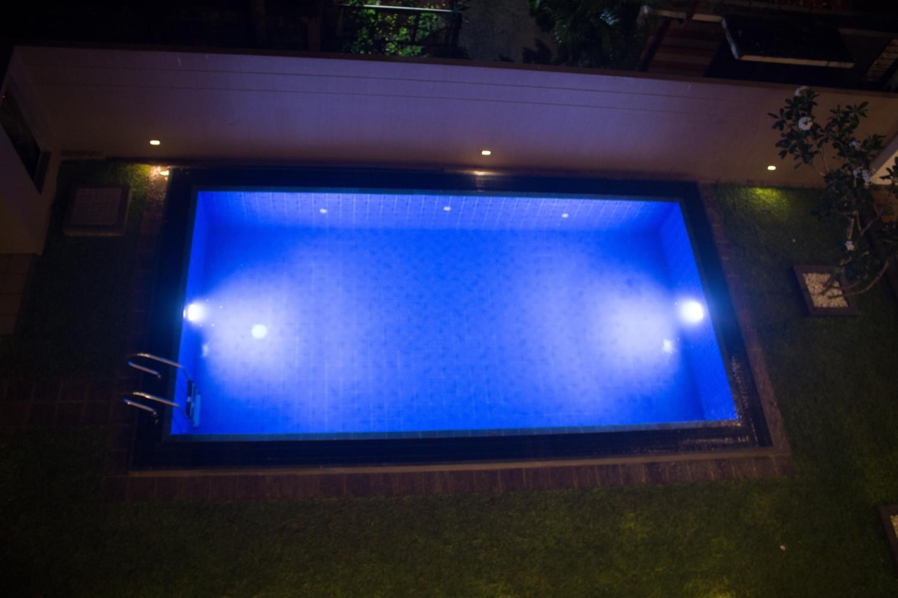 A rectangular swimming pool with a blue interior, surrounded by a brick border and illuminated by overhead lights.