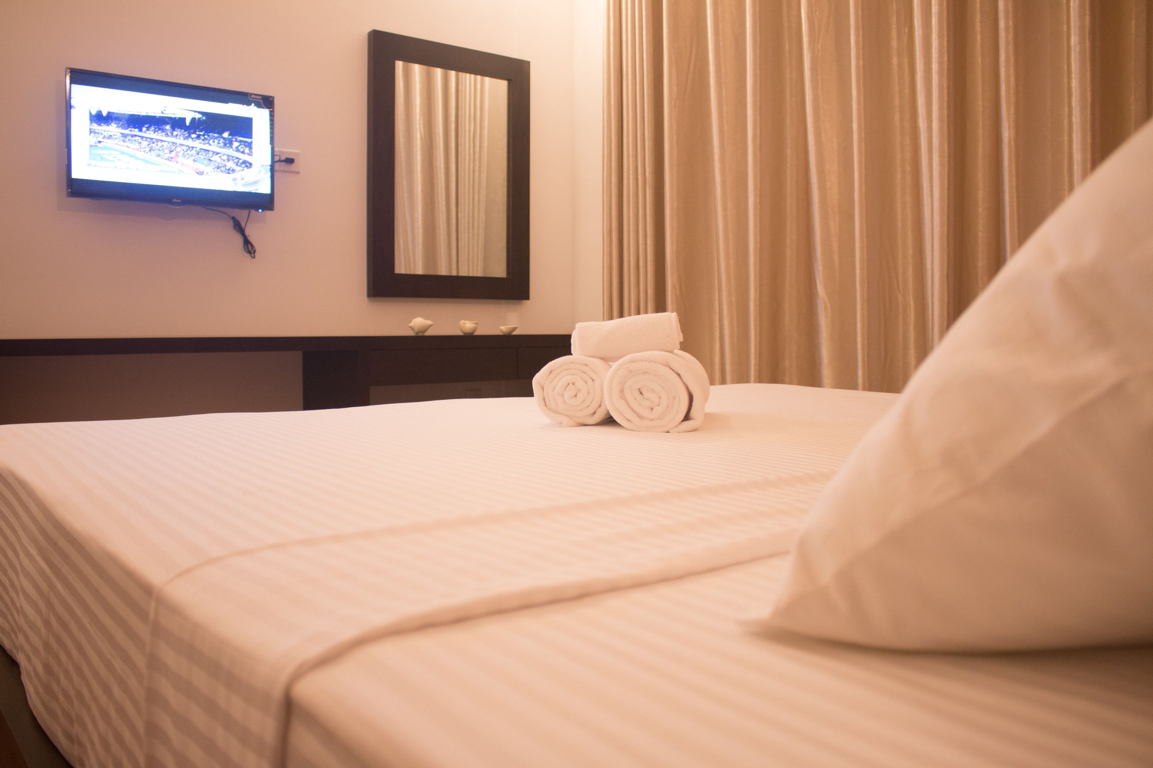 A neatly made bed with rolled towels and a pillow, a television mounted on the wall, and a mirror above it.