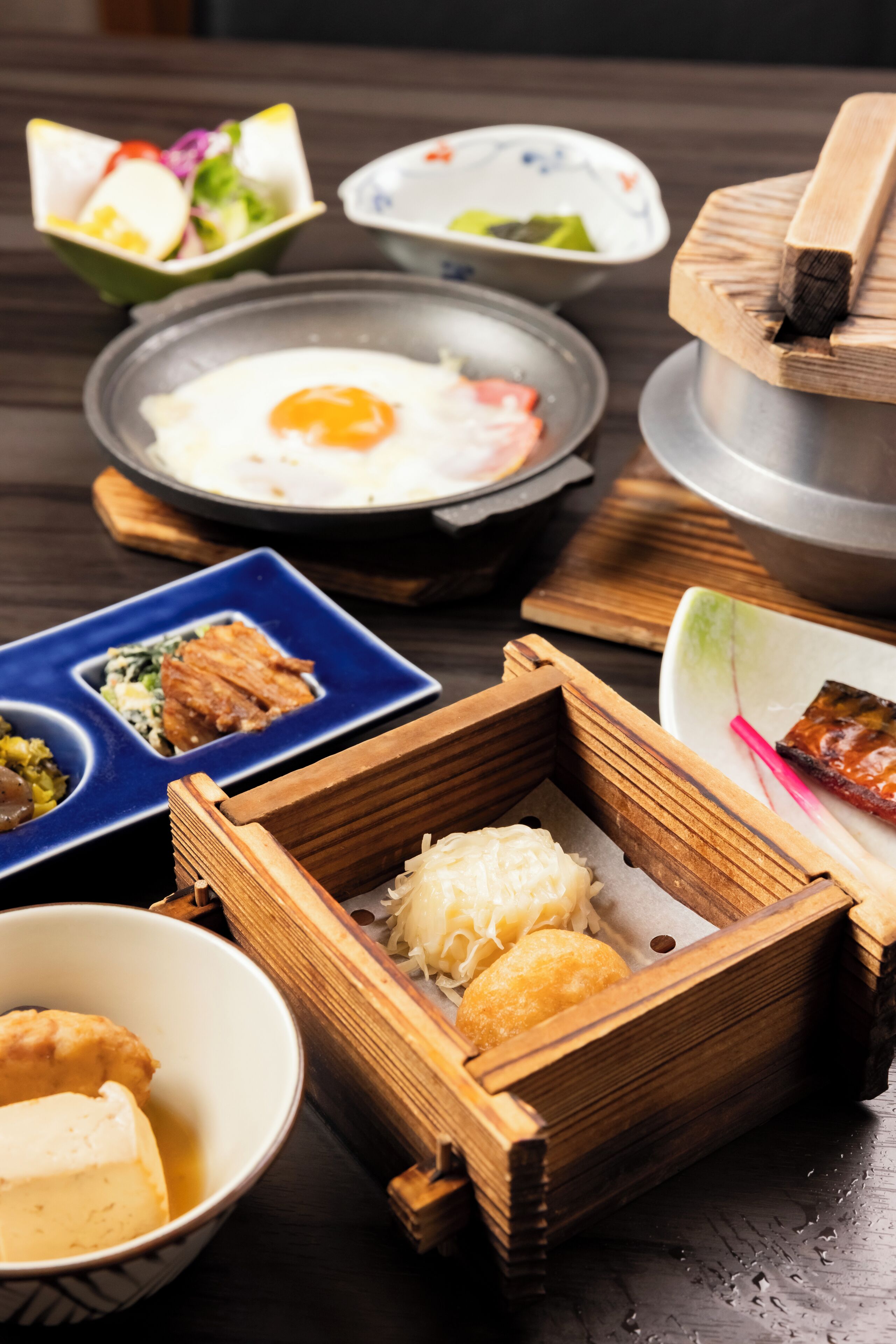A variety of dishes in traditional serving dishes, including a bowl of soup, a plate with a fried egg, and a wooden box with dumplings.