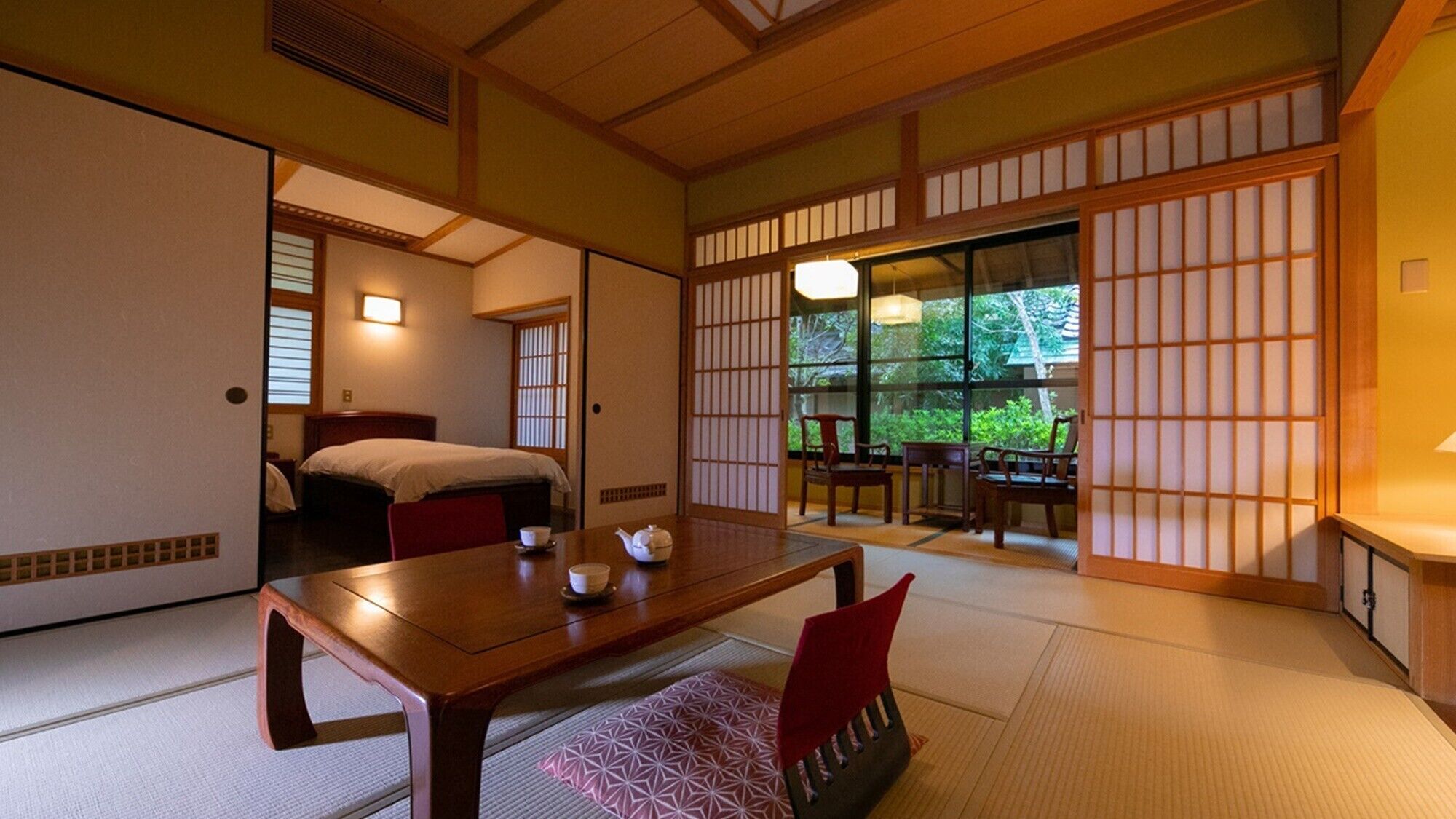 Een traditionele Japanse kamer met tatami vloerbedekking, een lage houten tafel met een theepot, een bed met futon, en schuifdeuren die uitkomen op een balkon met uitzicht op groen.