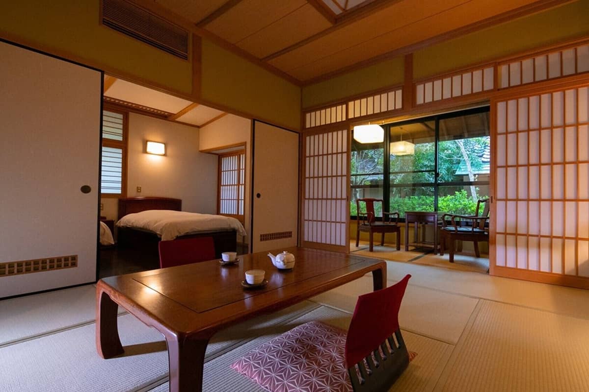 Una habitación japonesa tradicional con suelo de tatami, una mesa baja de madera con un hervidor, una cama con futón y puertas correderas que dan a un balcón con vistas a la vegetación.