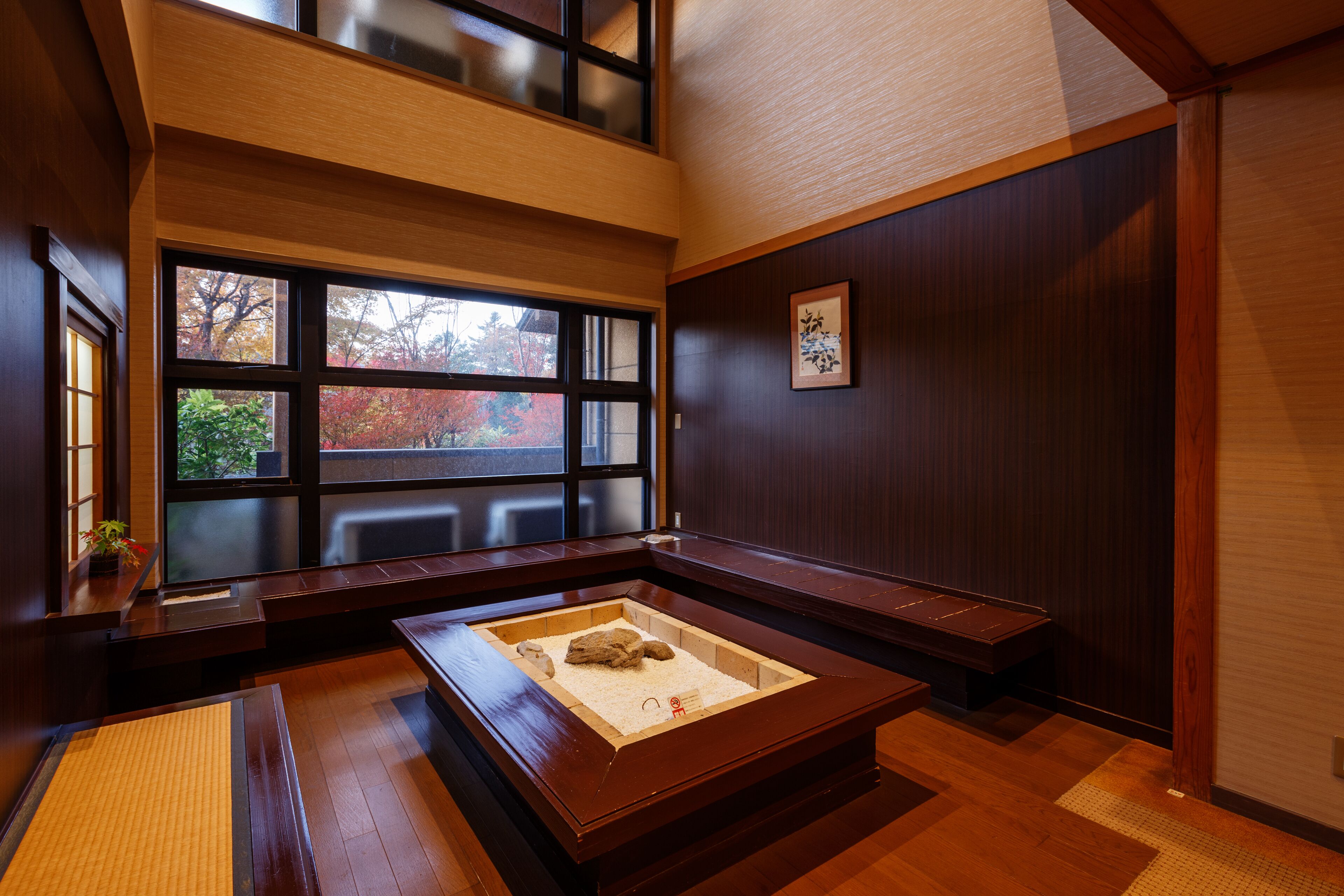 Una habitación japonesa tradicional con suelo de tatami, una mesa de madera baja con una composición rocosa en el centro y un ventanal con vistas a follaje otoñal.