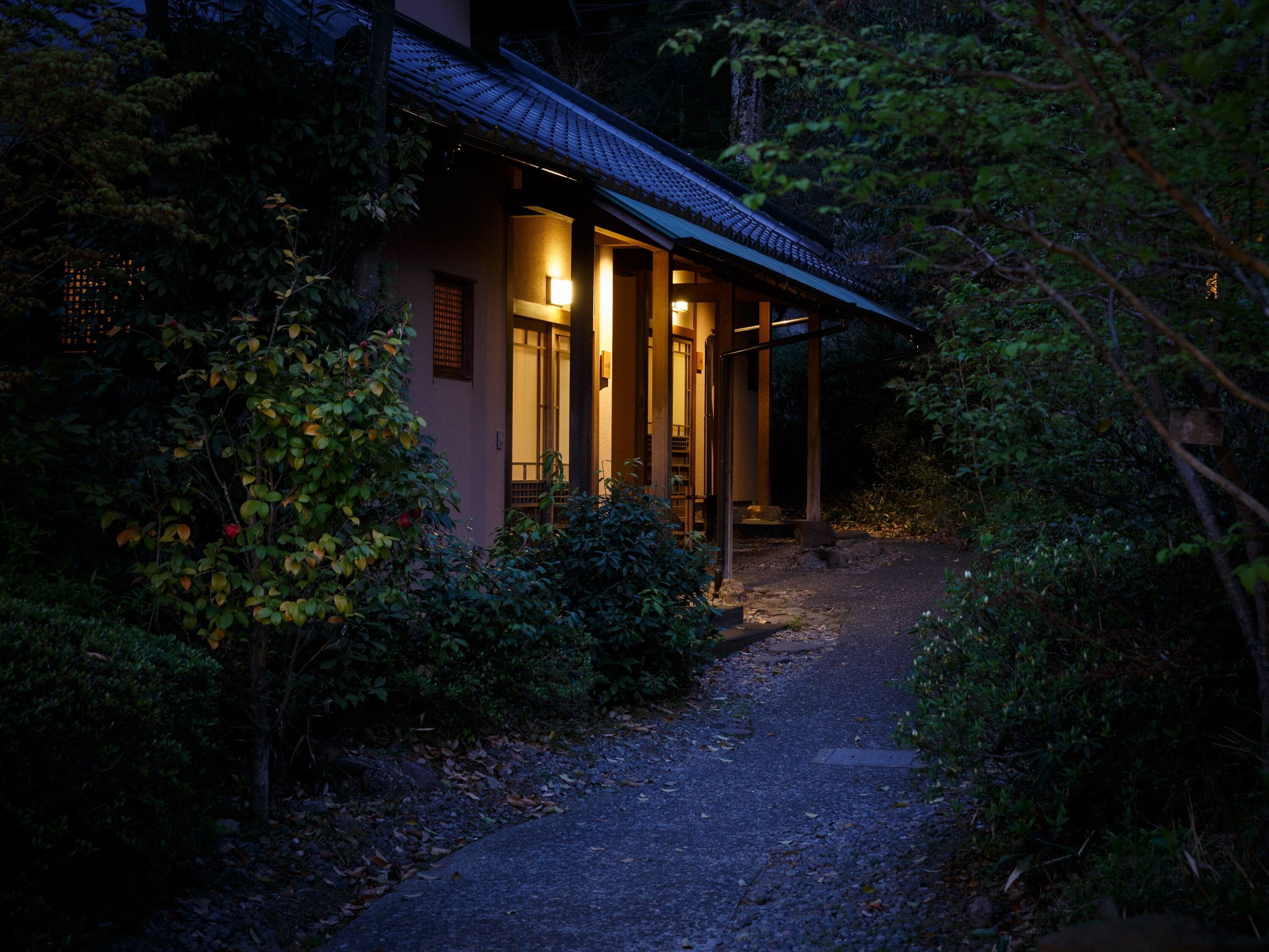 A traditional Japanese house with a gravel path leading to the entrance, surrounded by dense foliage.