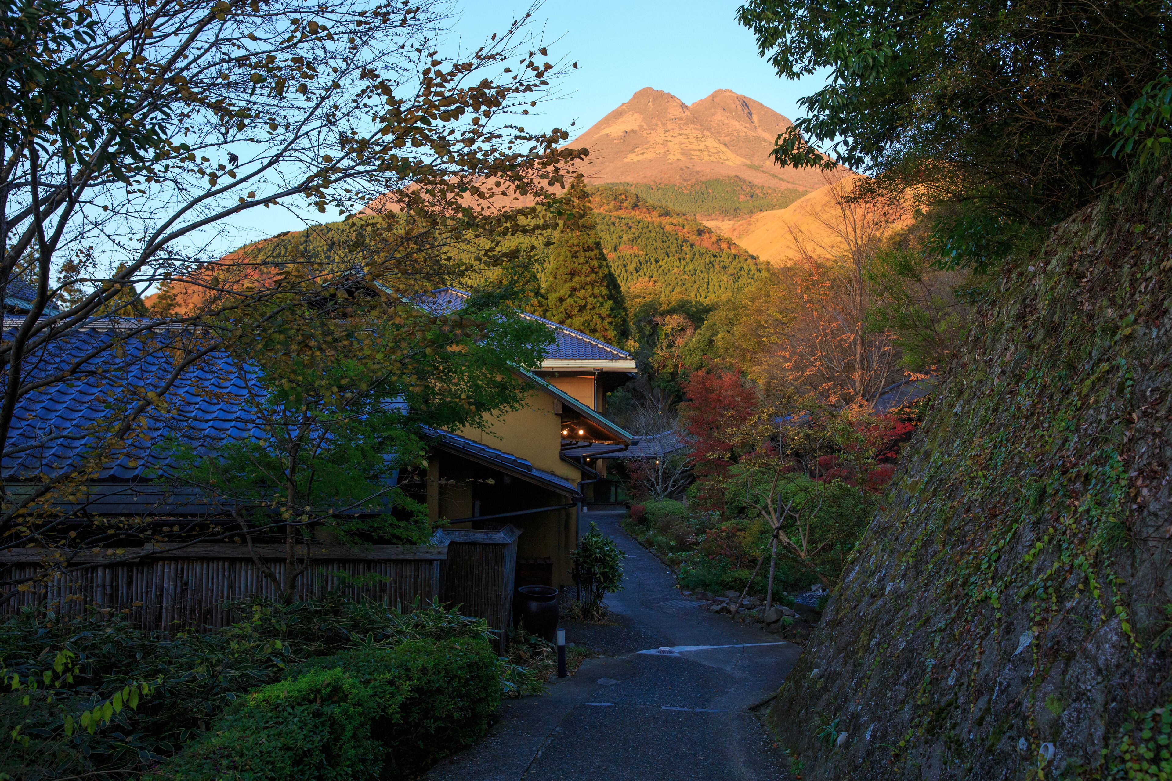 A narrow street leading to a traditional building with a sloped roof, surrounded by lush greenery and a mountain in the background.