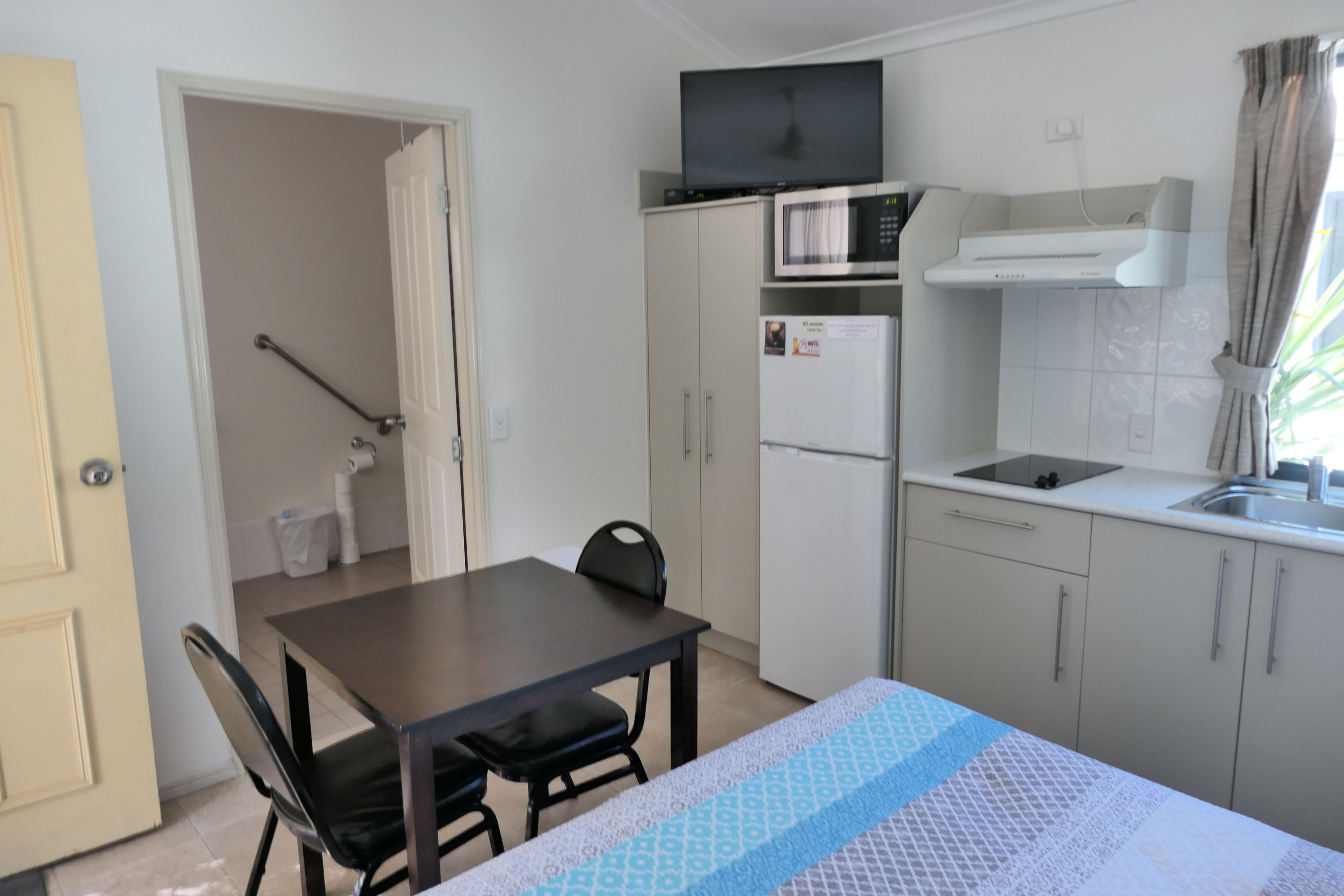 Een compacte keuken met een magnetron, koelkast en een klein spoelbakje. Er staat een eettafel met stoelen en een tafelkleed met een patroon.