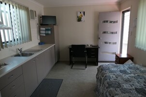 Habitación de hotel pequeña con kitchenette, una cama individual, un escritorio con silla, televisión y una puerta con número.
