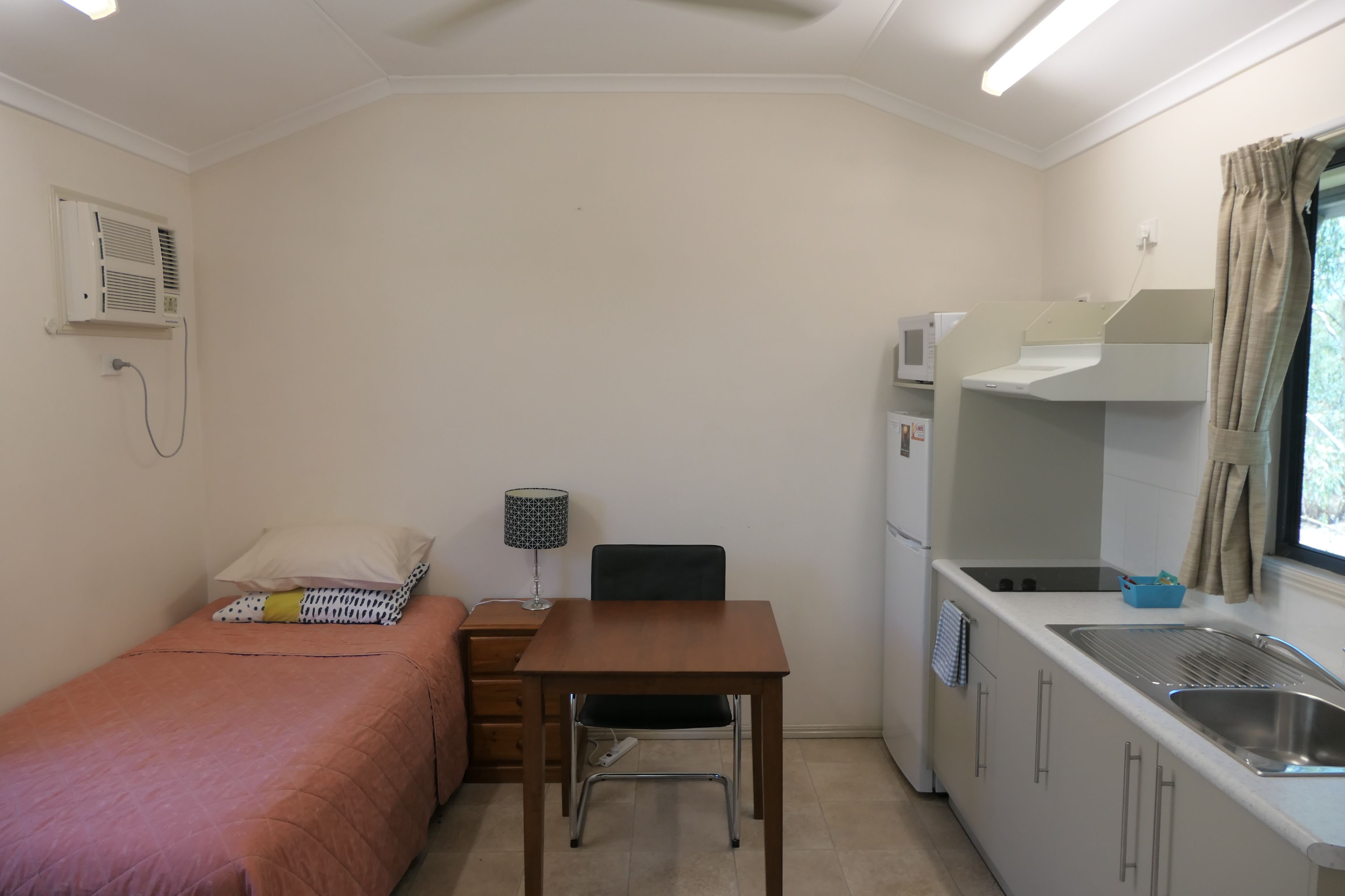 Une pièce de taille modeste comprenant un lit, une petite table, une chaise, un bureau, un four à micro-ondes, un évier et une fenêtre avec des rideaux.
