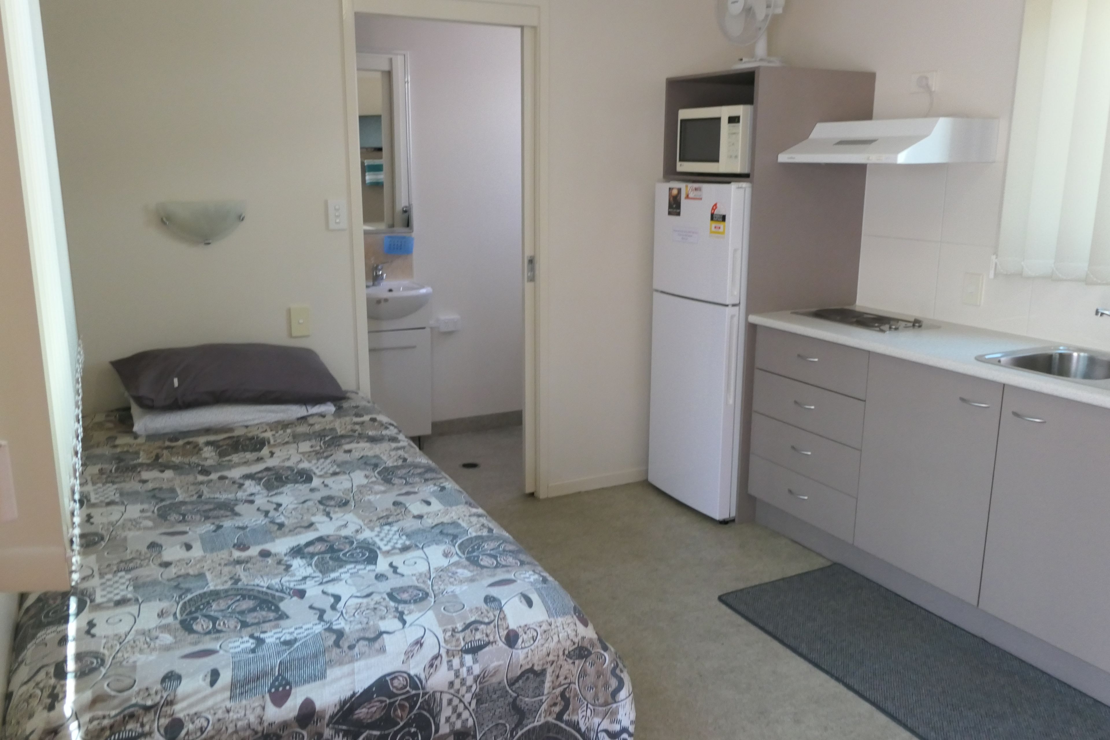 A small, well-lit room with a bed, a sink, a microwave, and a kitchenette.