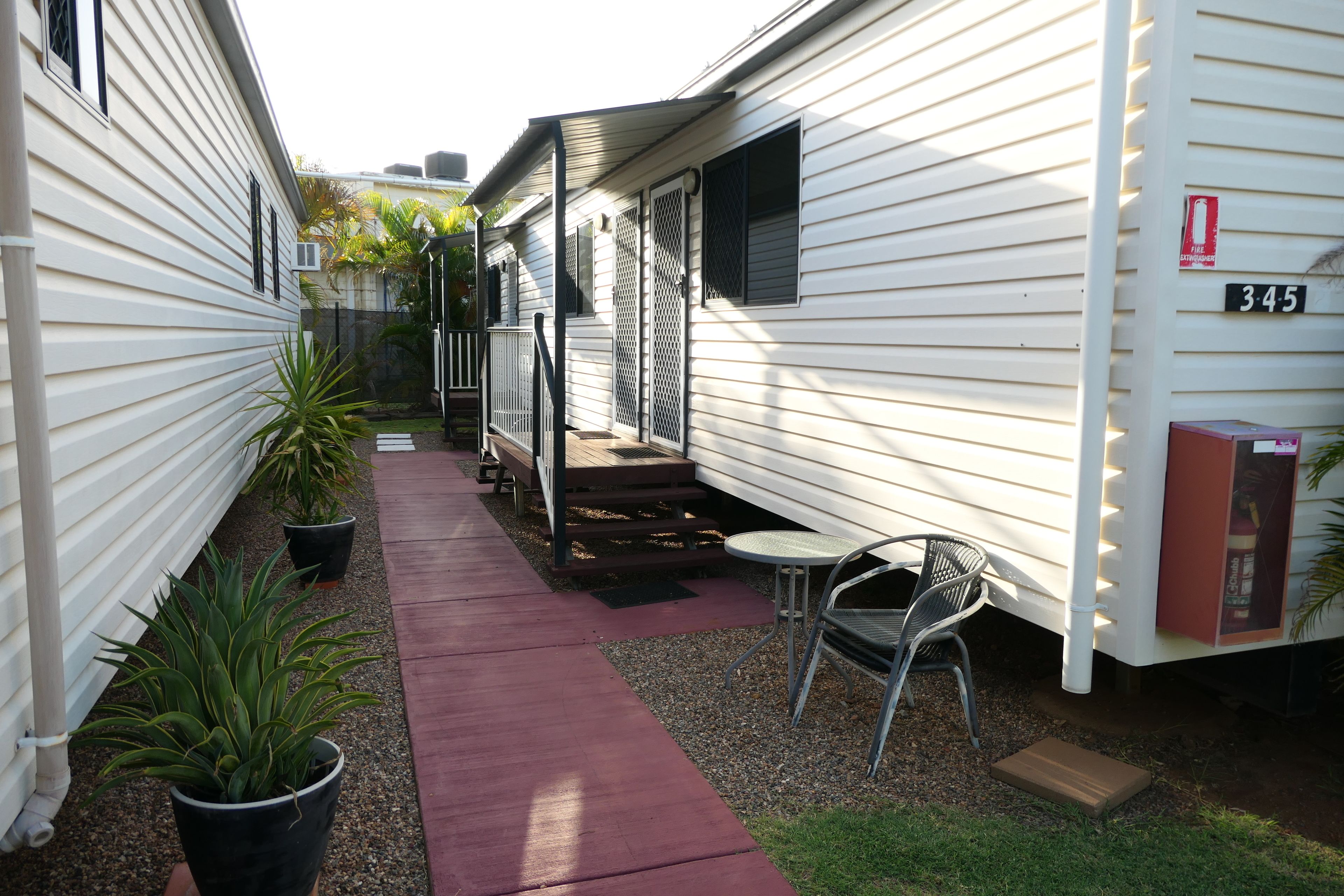 A narrow pathway between mobile homes with a red walkway, potted plants, and outdoor seating.