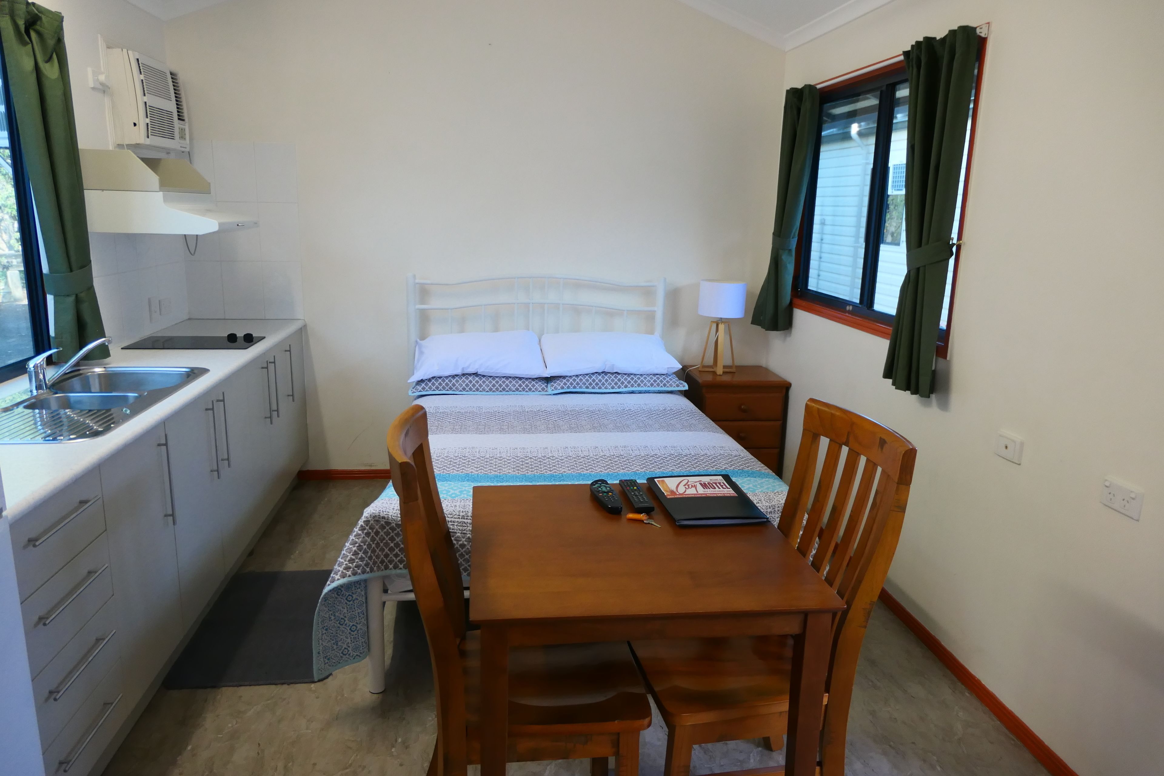 Une chambre d’hôtel compacte comprenant un coin cuisine, un lit avec une couvre-lit à motifs, une table en bois avec des chaises et une fenêtre avec des rideaux.