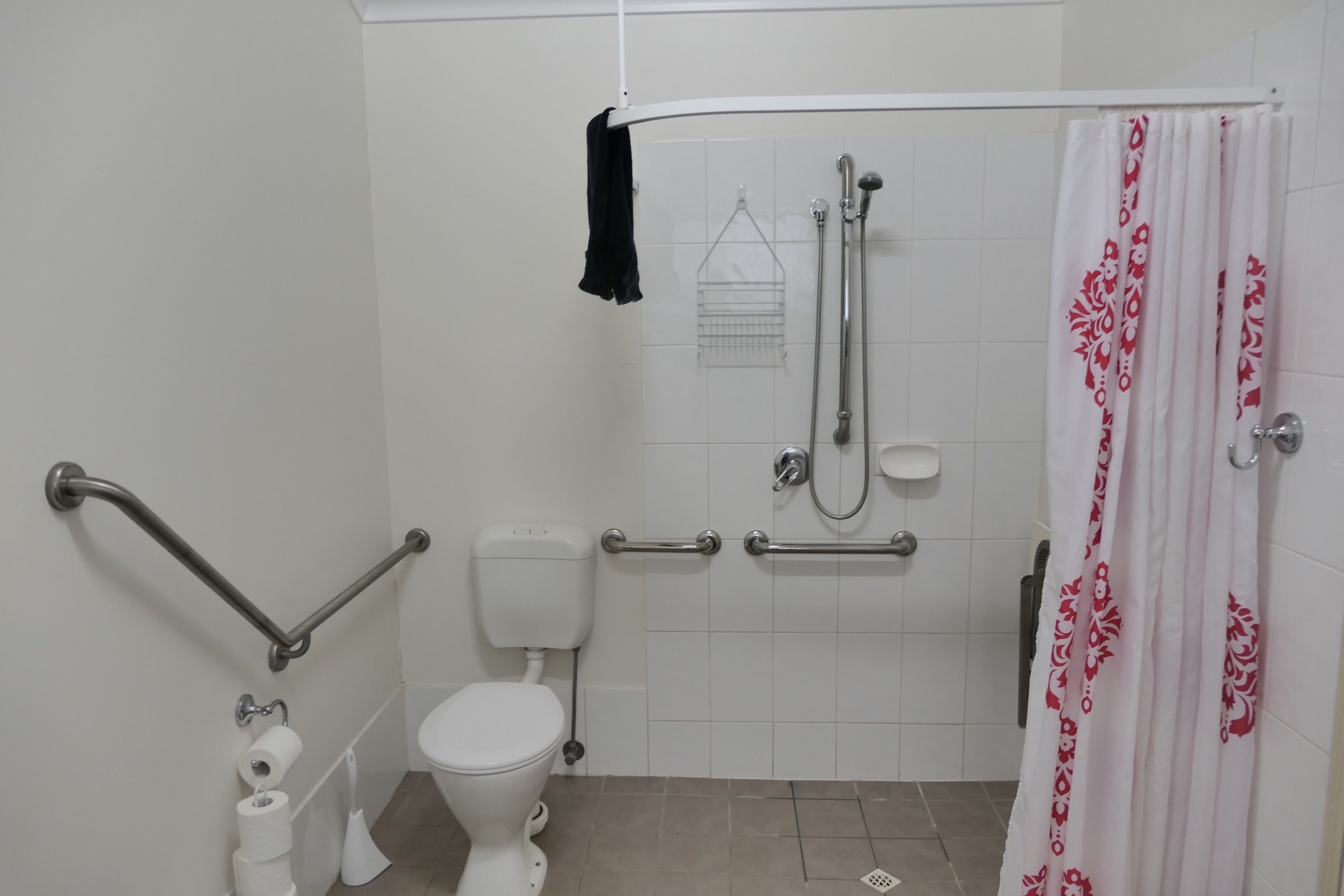 Une salle de bain équipée de toilettes, de barres d’appui, d’une douche et d’un mur recouvert de carreaux blancs.