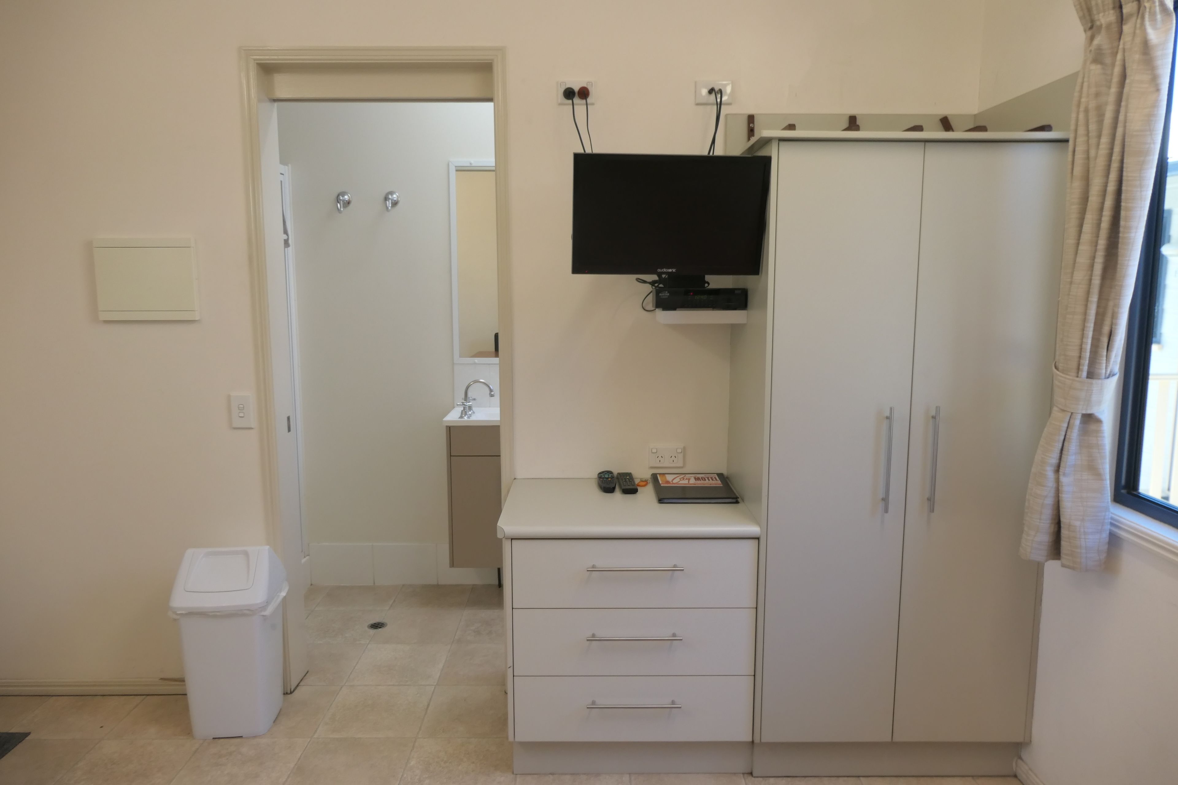 Habitación pequeña y ordenada con un armario blanco, un televisor de pantalla plana montado encima, un escritorio pequeño con una computadora y una ventana con cortinas.