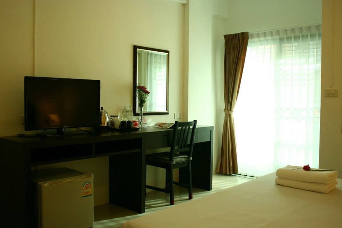 Habitación de hotel con cama, escritorio con televisor, silla, refrigerador pequeño, espejo y ventana con cortinas.