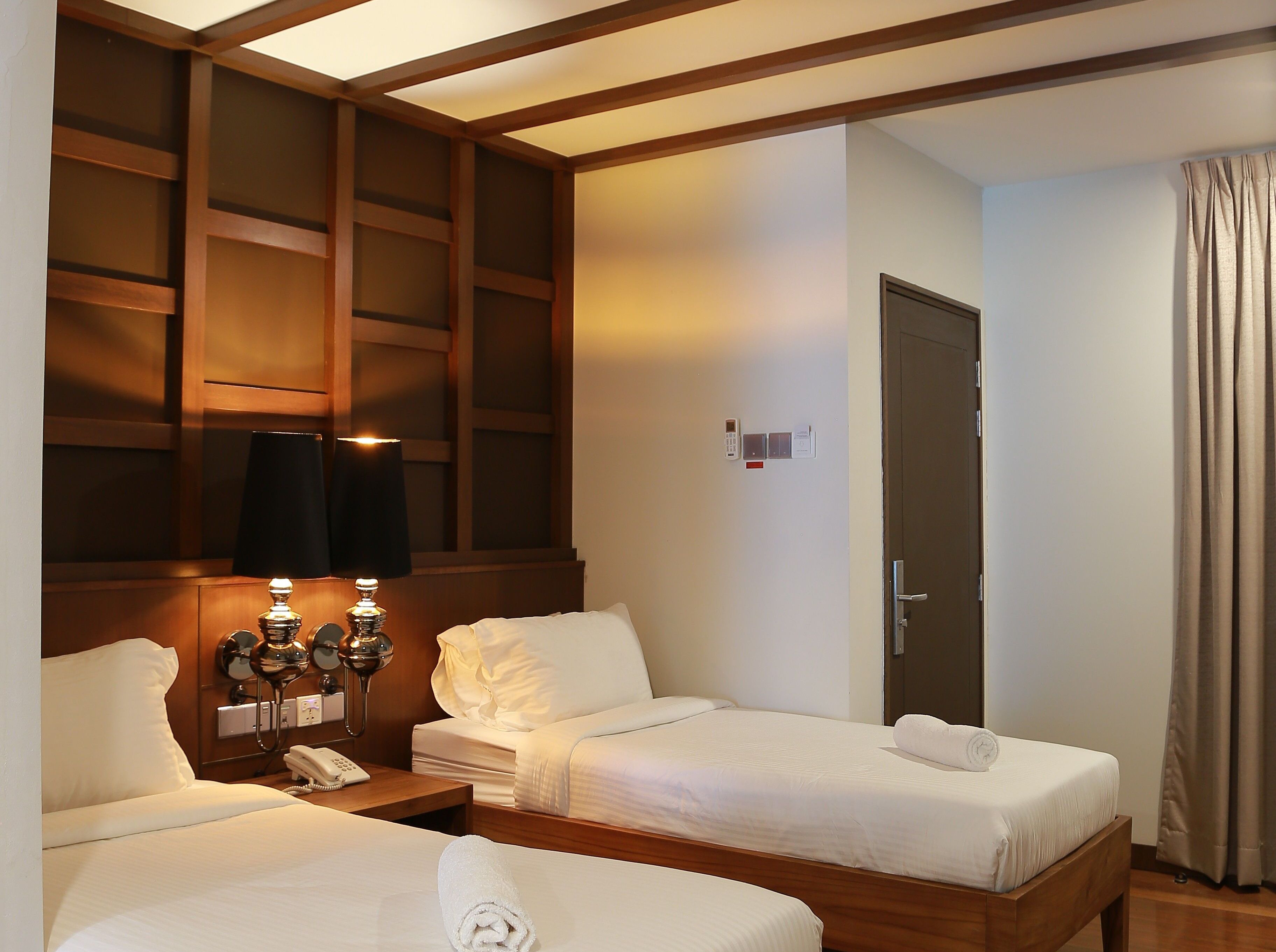 A hotel room with two beds, a wooden headboard, a bedside table with a lamp, a phone, and a door with a lock.