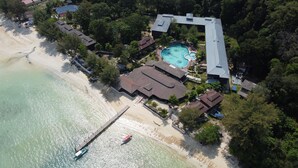 Vista aérea de un complejo turístico con playa, piscina y un muelle con un bote.