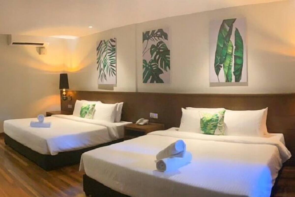 Zwei Doppelbetten mit weißen Bettwäsche und Kissen mit grünem Blattmuster, Holzboden und eine Wandlampe.