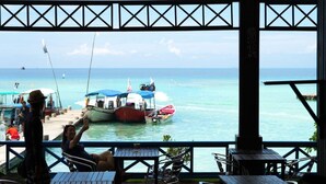 Un restaurante costero con terraza al mar, con mesas y sillas, y barcos amarrados cerca.