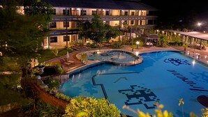 Una piscina con diseño geométrico, rodeada de zonas ajardinadas con árboles y un edificio con varios balcones al fondo.