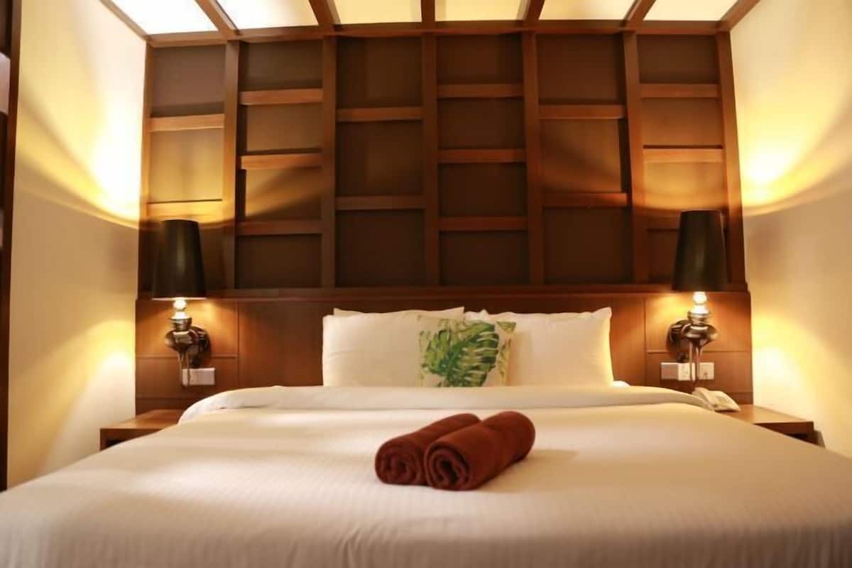 Ein Hotelzimmer mit Holzkopfteil, zwei Nachttischlampen, einem ordentlich bezogenen Bett und einem aufgerollten Handtuch auf dem Bett.