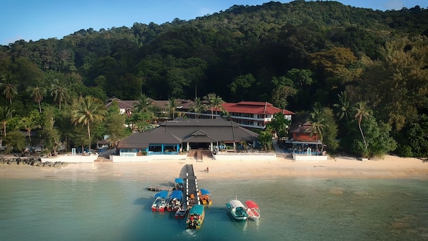 Un hotel frente al mar, con playa, barcos y una colina boscosa al fondo.