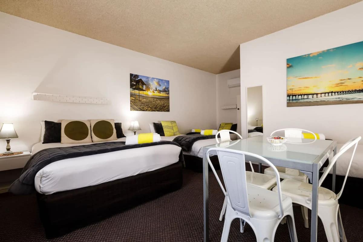 A hotel room with two beds, a dining table with chairs, a mirror, and two paintings on the walls.