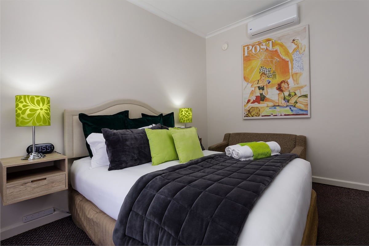 A neatly made bed with a dark quilt, two green pillows, and a white comforter. There is a bedside table with a lamp and a digital clock. A poster of a beach scene is on the wall.