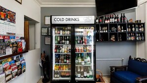 Una habitación con un refrigerador que indica 'BEBIDAS FRÍAS', repleto de bebidas, una estantería con varios objetos y un sofá azul.