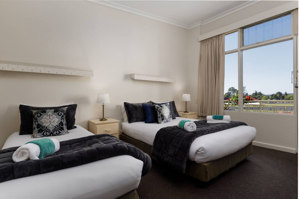 A hotel room with two beds, each with a black and white patterned pillow, a white bedside table with a lamp, and a window with a view of a park and buildings.