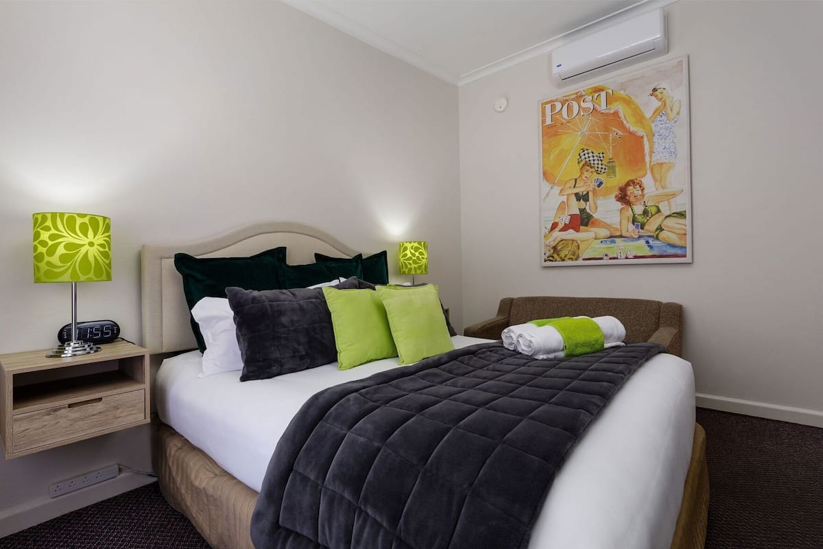 A hotel room with a bed, a nightstand with a lamp, a wall clock, and a poster.