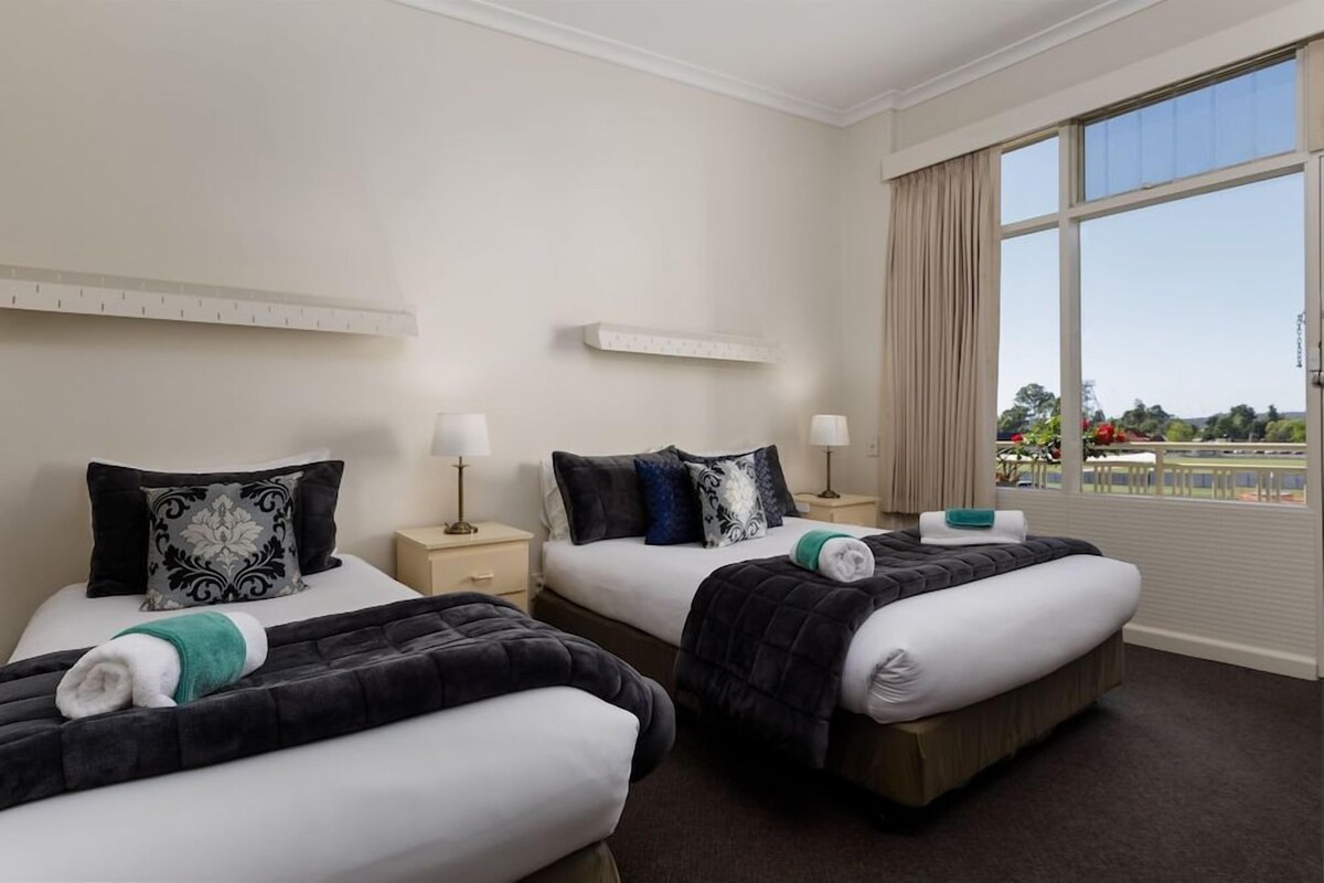 A hotel room with two beds, each with a pillow, a bedside table with a lamp, and a window with curtains.