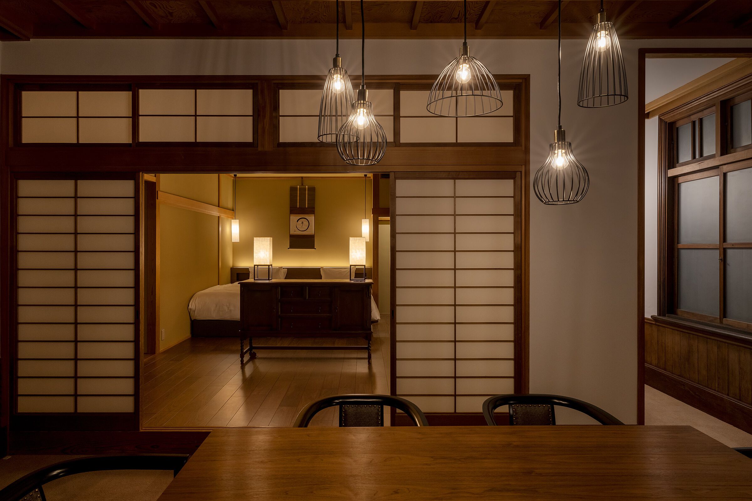 A traditional Japanese room with tatami flooring, a low table, and a bed with a futon. There are three hanging light fixtures and a sliding shoji screen door.