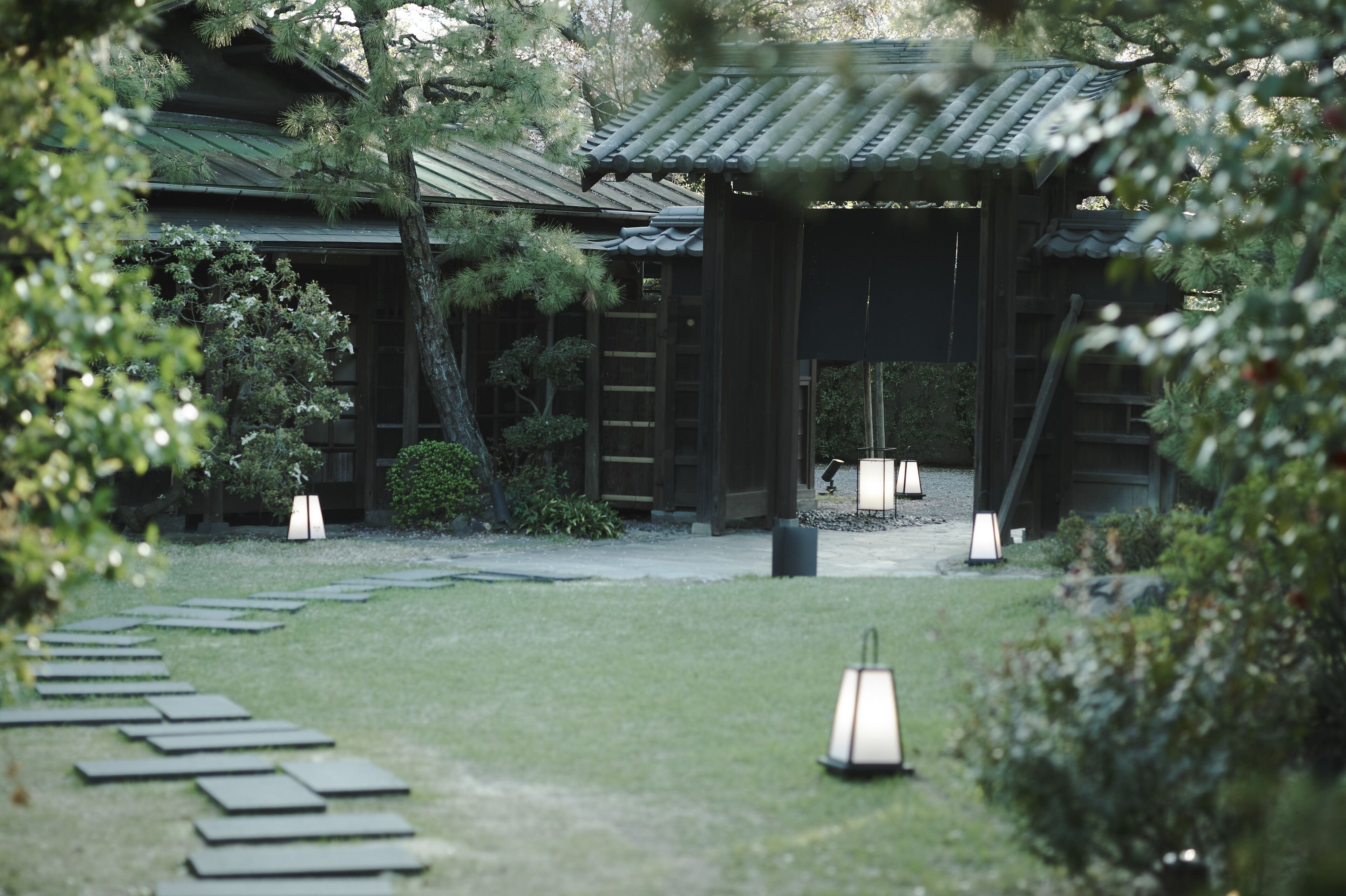 A traditional Japanese-style building with a wooden structure and tiled roof, surrounded by a well-maintained garden with stone lanterns and a stone pathway.
