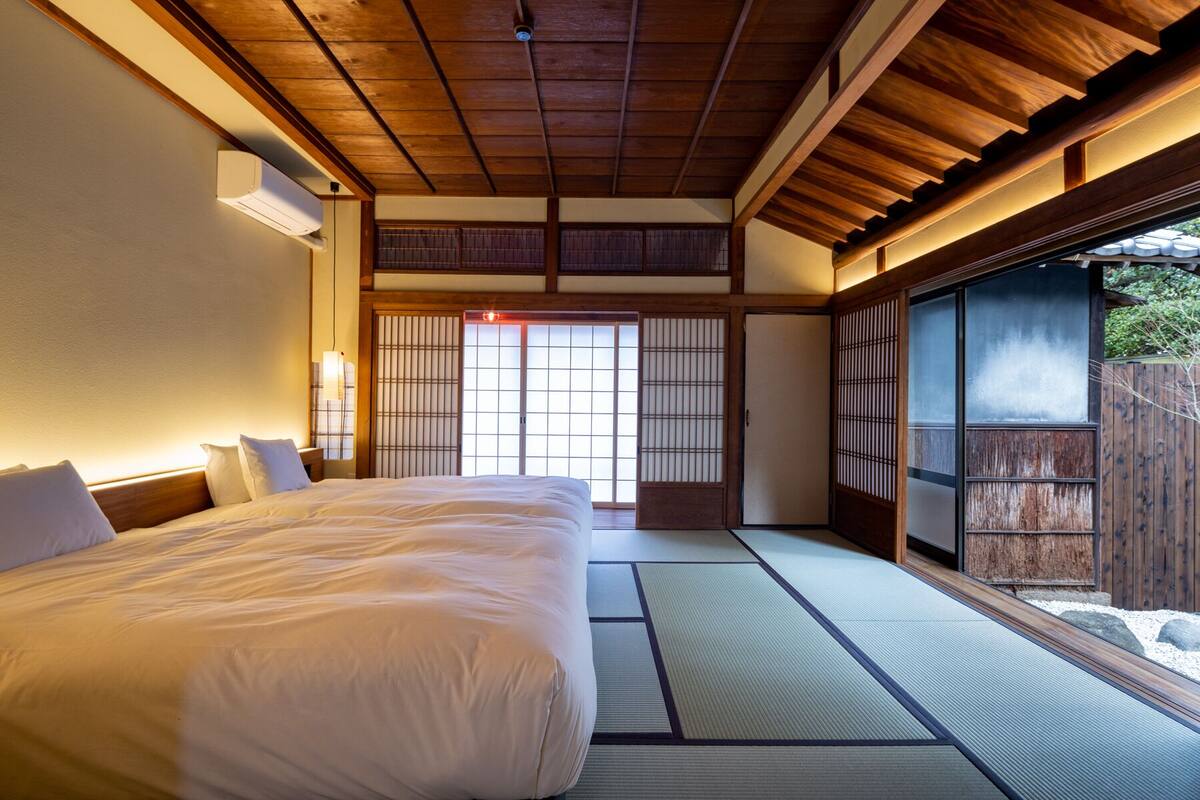 Un dormitorio de estilo japonés tradicional con suelo de tatami, una cama grande, puertas correderas shoji y vistas a un jardín.