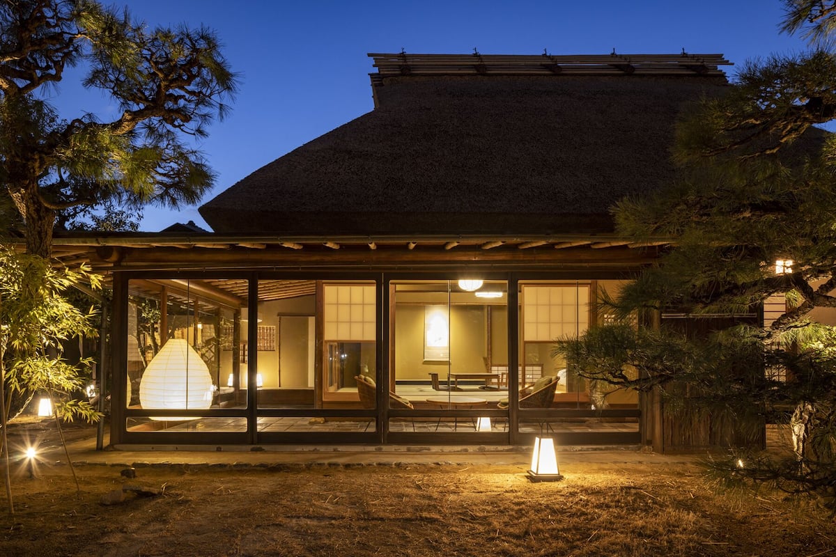 Un edificio de estilo japonés tradicional con techo de paja, amplios ventanales correderos y un área de estar al aire libre techada.