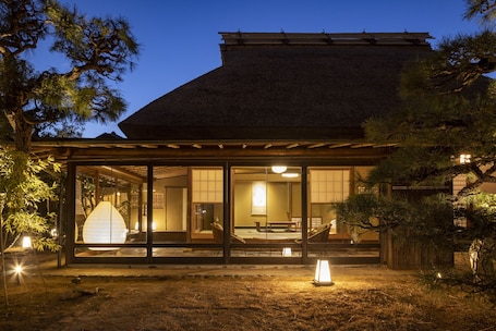 A traditional Japanese-style building with a thatched roof, large sliding glass doors, and a covered outdoor seating area.