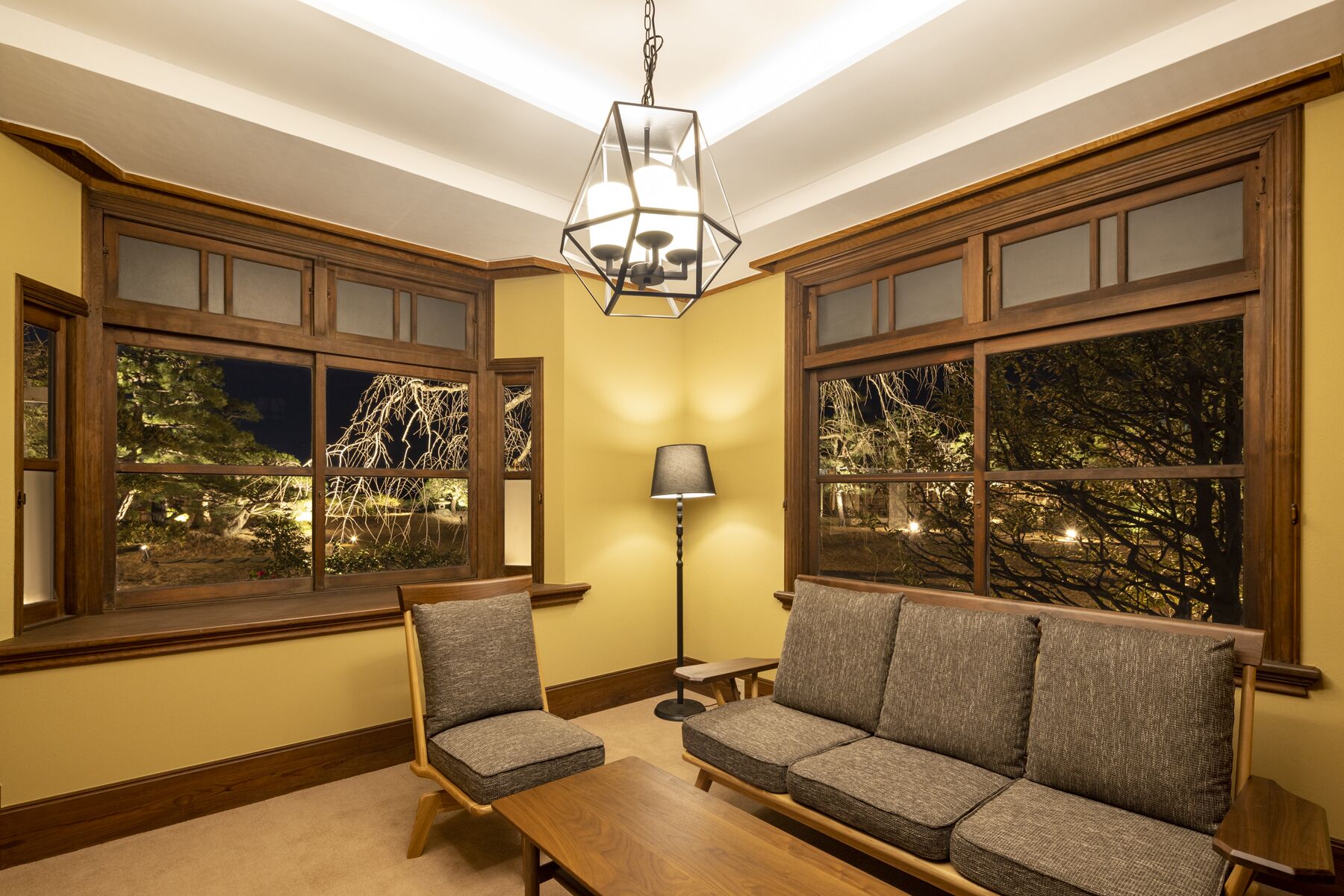 A cozy room with a wooden bench, a cushioned chair, and a sofa facing large windows with a view of trees.
