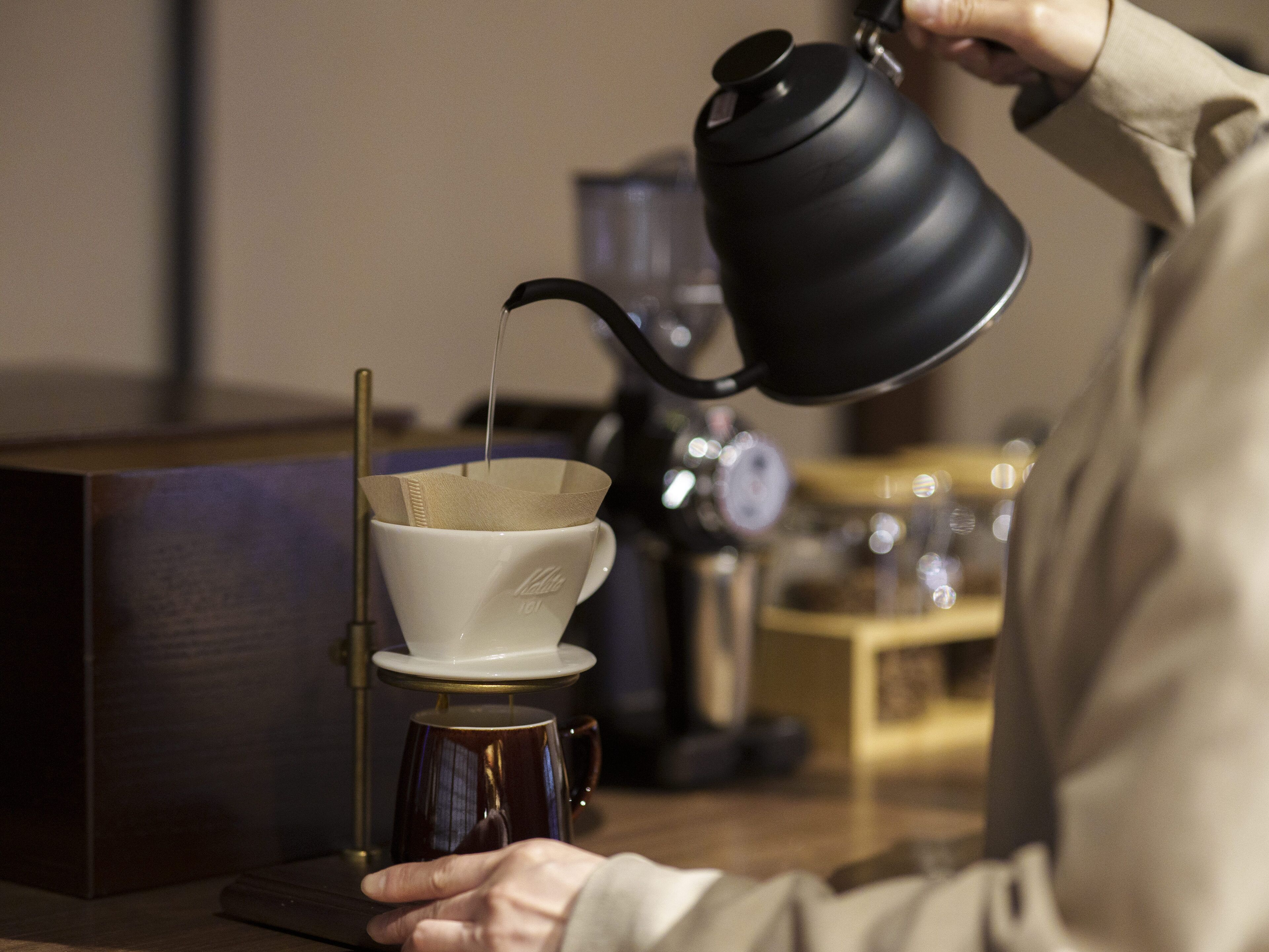 A person pouring liquid from a black kettle into a white cup with a paper filter.