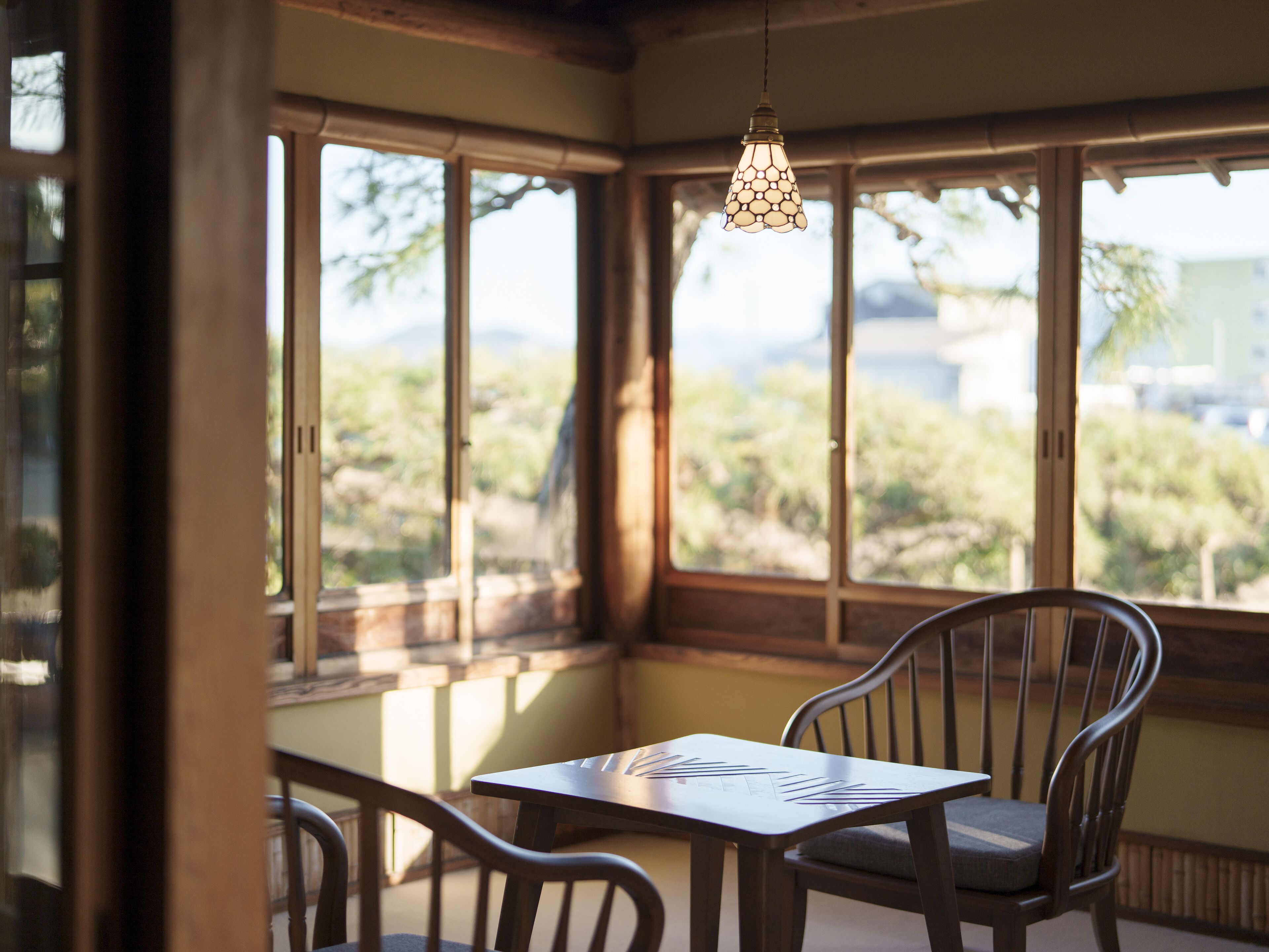 A room with a table and chairs, large windows with a view of greenery, and a hanging light fixture.