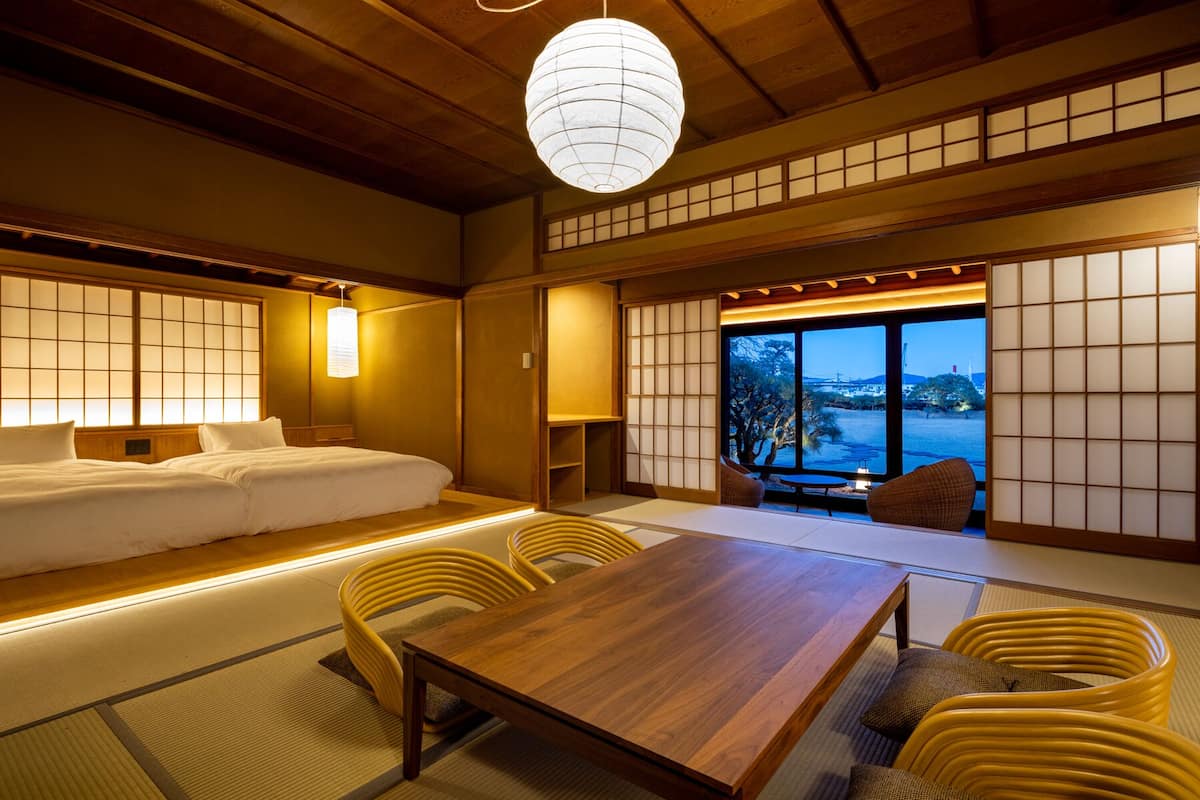 Una habitación japonesa tradicional con suelo de tatami, una mesa de madera baja, sillas tejidas amarillas, una cama con ropa de cama blanca y vistas a un cuerpo de agua a través de biombos shoji.