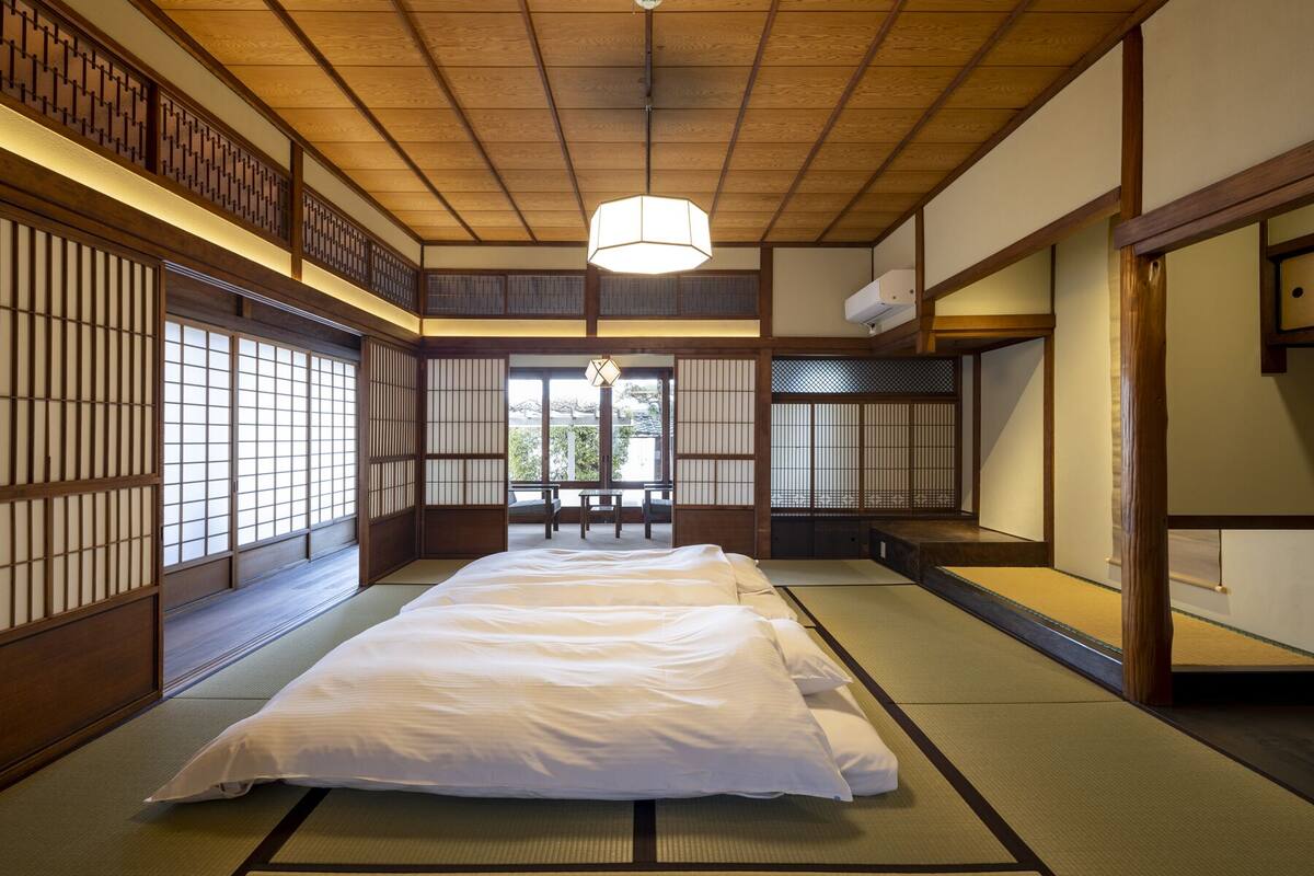 Una habitación japonesa tradicional con suelo de tatami, una cama baja, puertas correderas y una lámpara colgante.