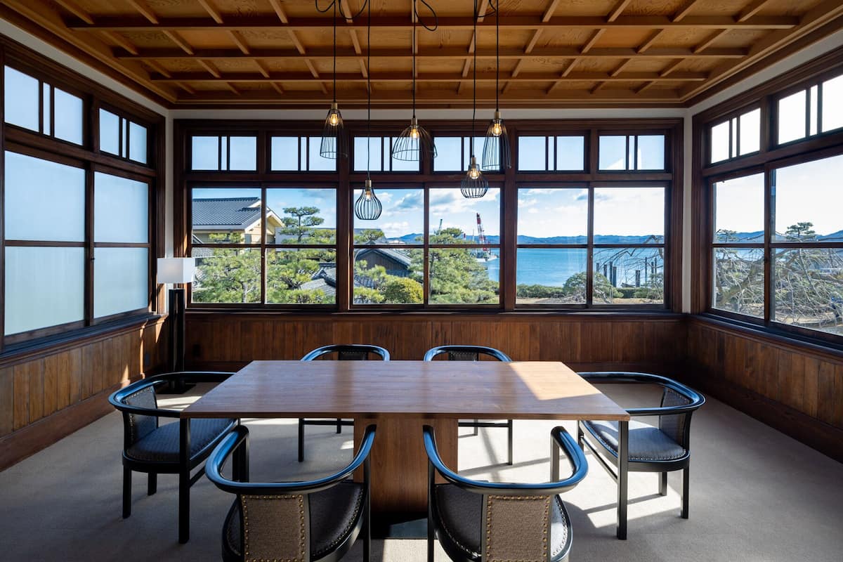 Un comedor con una mesa y sillas de madera, ventanas amplias con vistas a un paisaje costero.