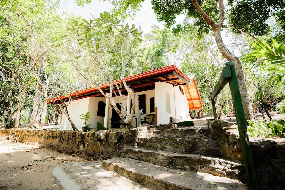 Un edificio rústico con techo rojo, rodeado de abundante vegetación y escalones de piedra que conducen a él.