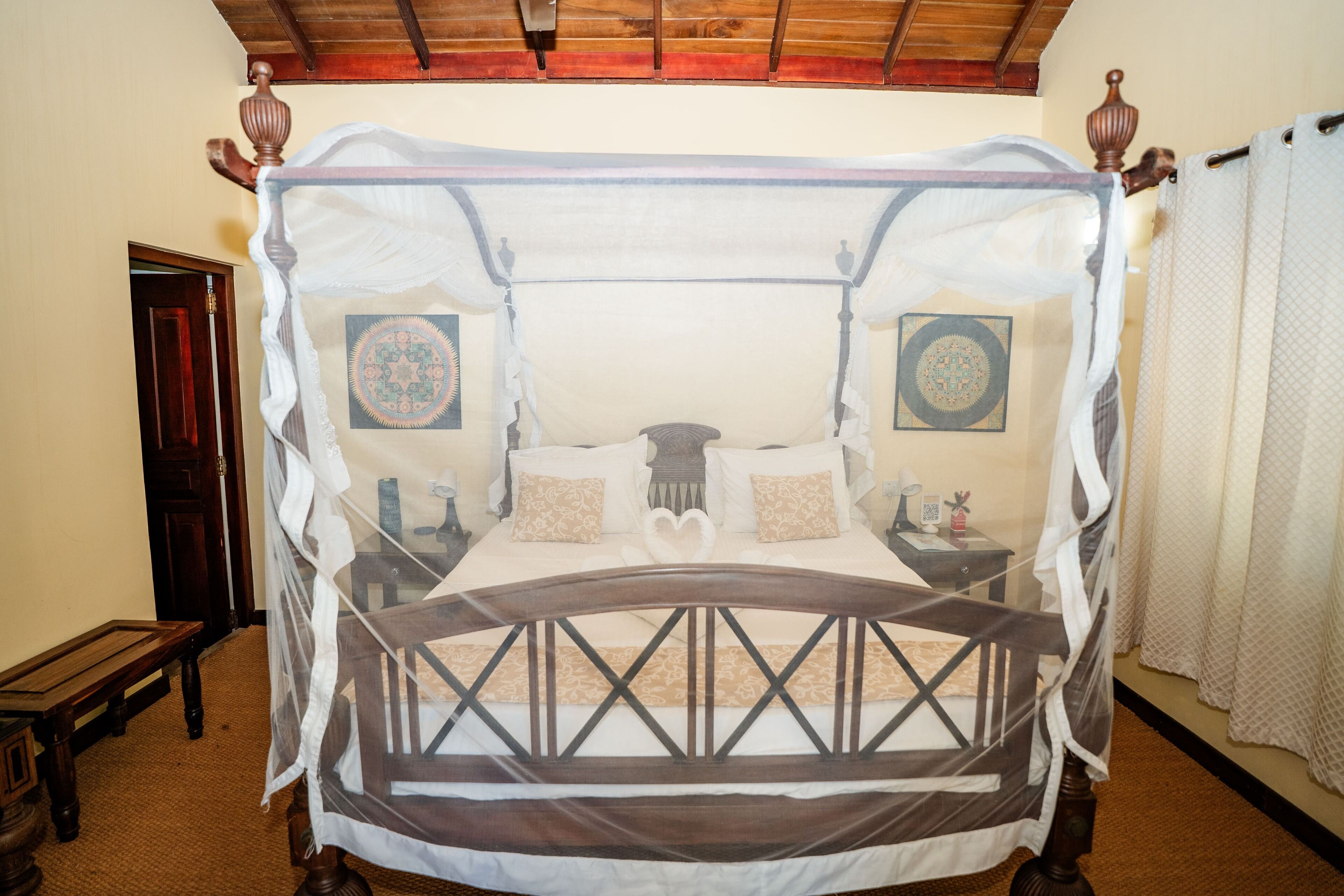 A four-poster canopy bed with a wooden frame and white bedding, two bedside tables with lamps, and a patterned wall hanging.