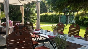 Un área de comedor al aire libre con sillas y mesas de madera, un espacio de estar techado y un jardín de fondo.