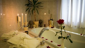Un baño lujoso con una bañera grande, toallas blancas, un jarrón con una rosa y objetos decorativos como velas y una planta.