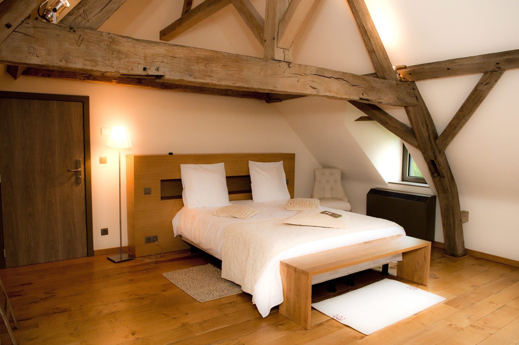 Ein Schlafzimmer mit Holzbalkendecke, einem Bett mit weißer Bettwäsche, einem Nachttisch, einer Lampe und einem Fenster.
