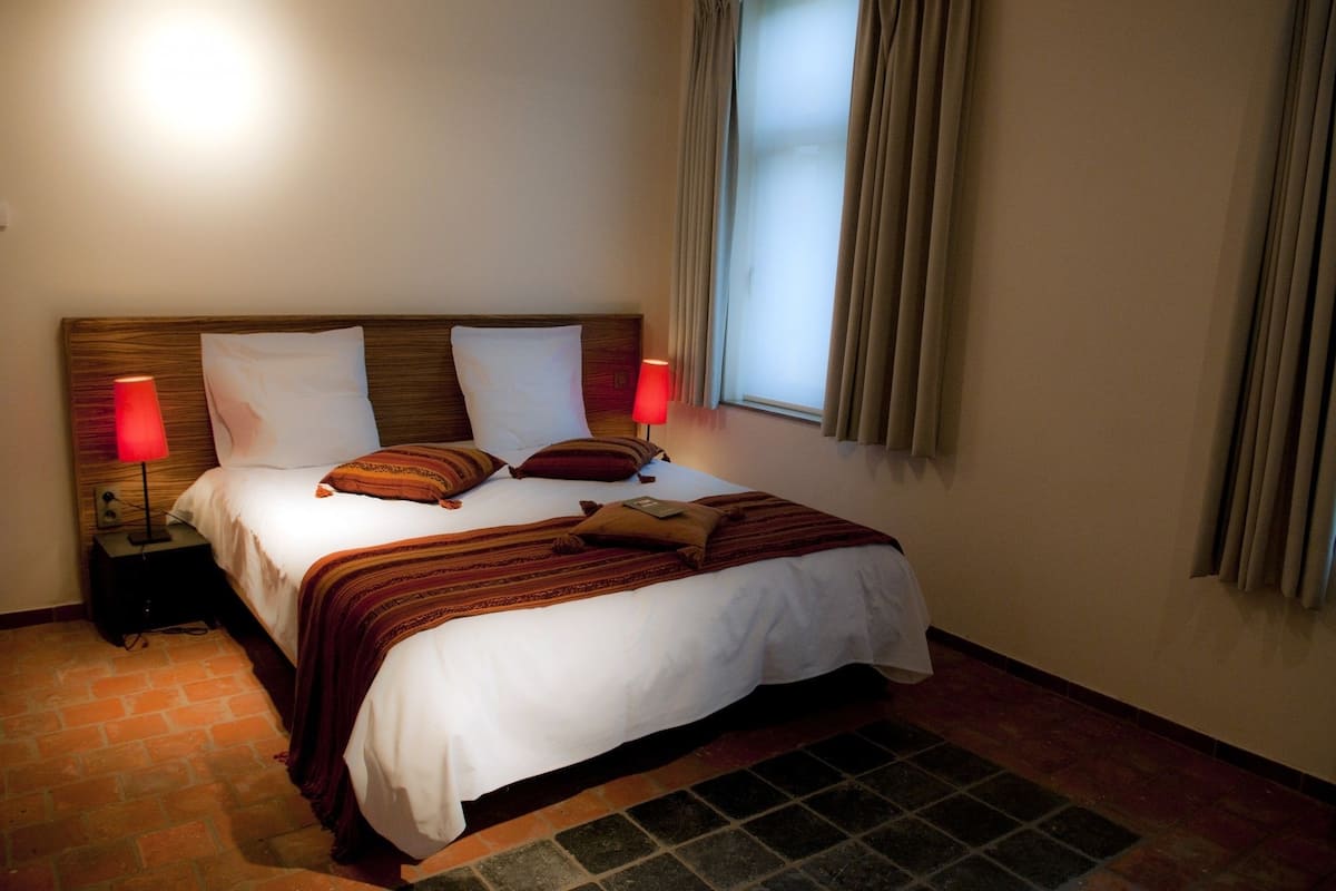 A hotel room with a wooden headboard, two bedside lamps, a bed with white and brown bedding, and a window with curtains.